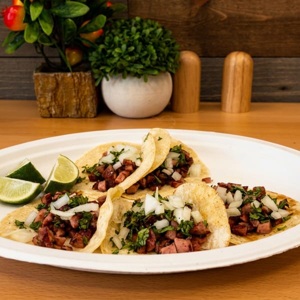 Three tacos filled with diced grilled meat, onions, and cilantro, served with lime wedges on a white plate.