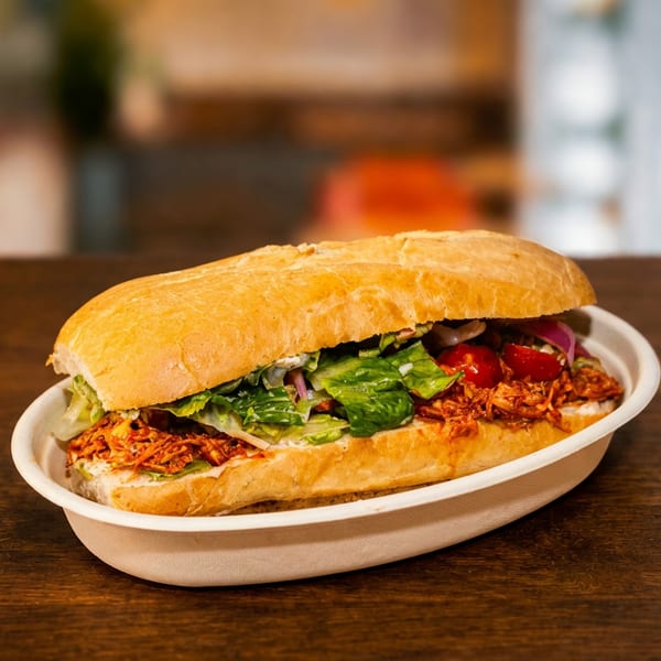 A loaded sandwich with lettuce, tomatoes, onions, and crispy shredded topping sits in a compostable tray on a wooden restaurant table.