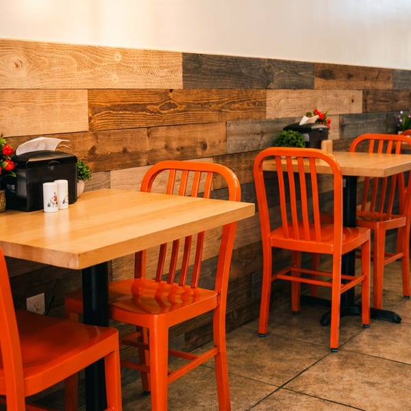 Restaurant dining area with wooden tables, bright orange chairs, floral centerpieces, and a rustic wood-paneled wall.