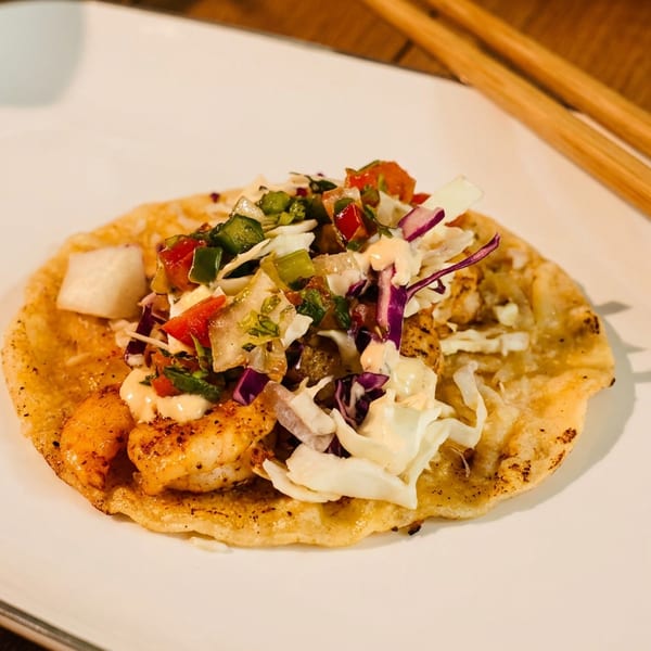 Shrimp tostada topped with cabbage, red onion, diced peppers, salsa, and creamy sauce on a white plate.