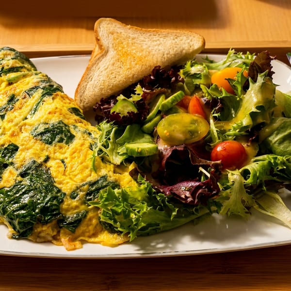 Spinach omelet served with mixed green salad, cherry tomatoes, toast, butter, and wrapped cutlery on a wooden tray.